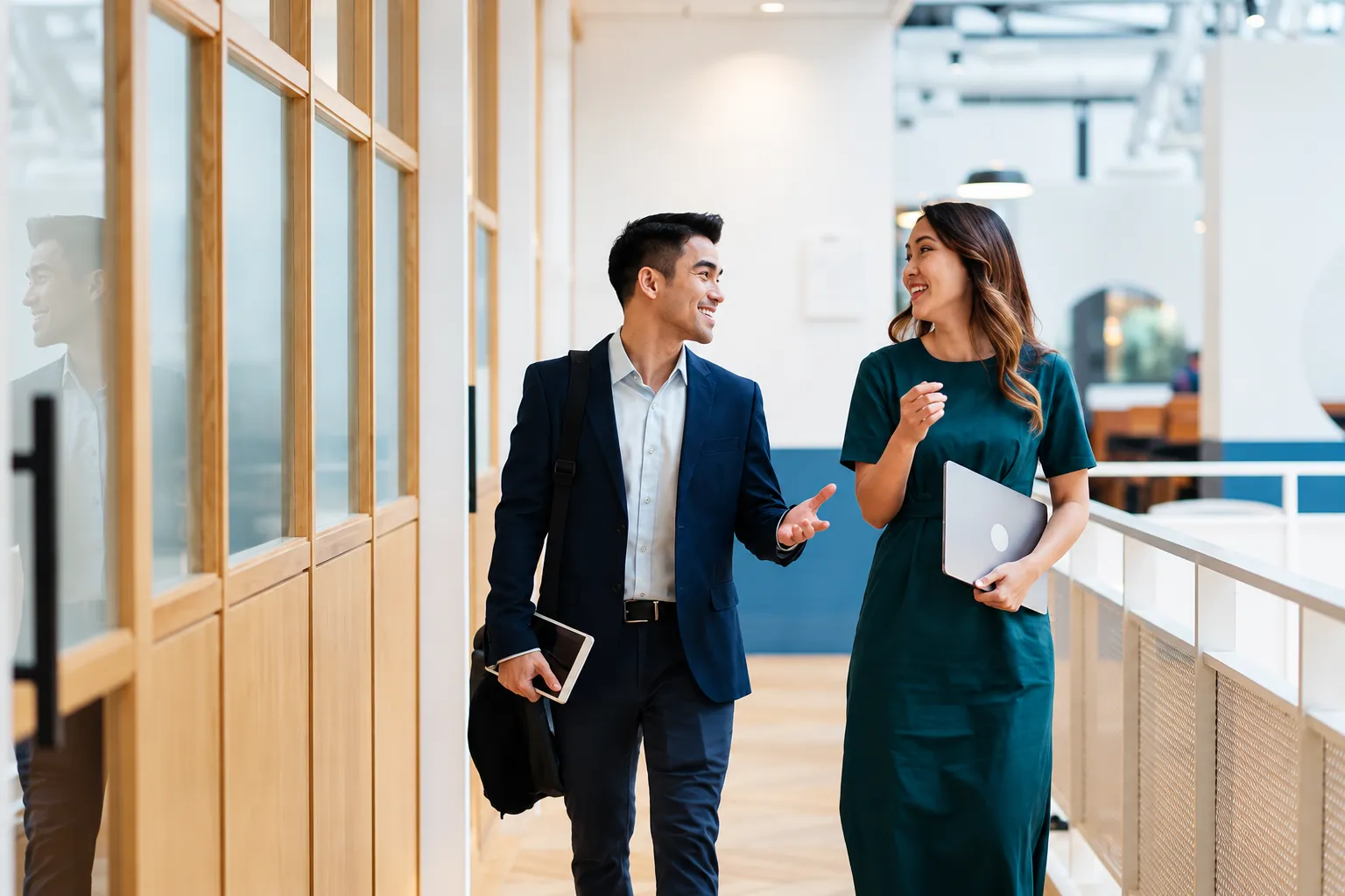 Two colleagues walking through a bright office corridor with a laptop and notebook