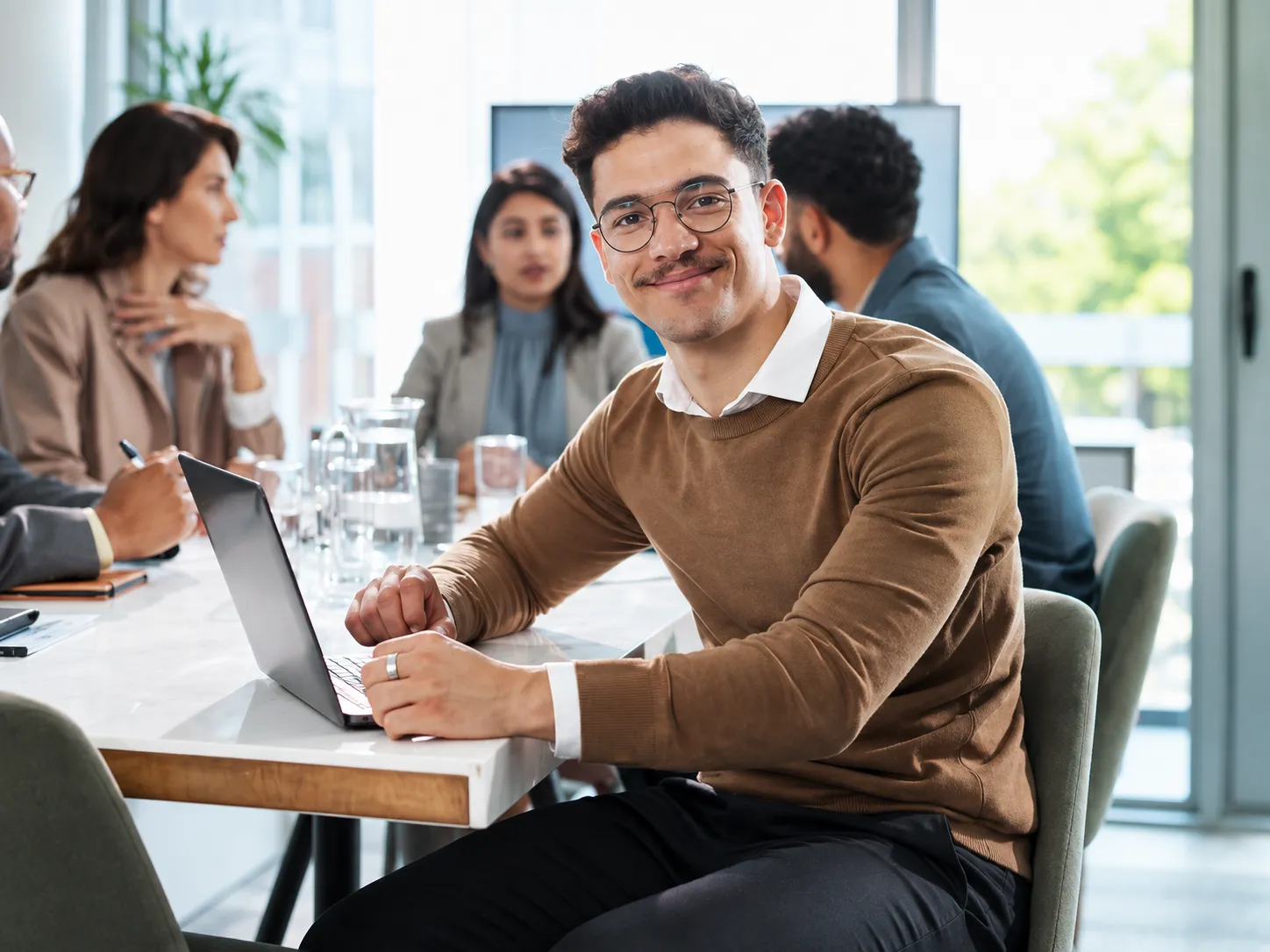 A smiling professional working on a laptop with colleagues in a bright office